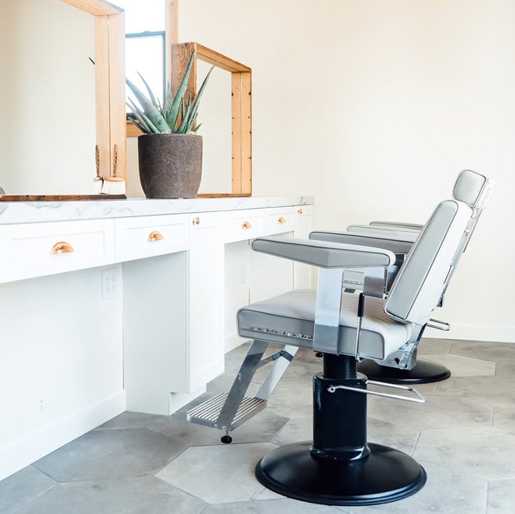 Side view of a Takara Belmont Dainty barber chair with light grey upholstery at a grooming station